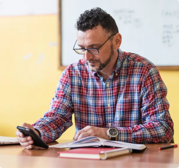 A man with glasses examines his phone while seated at a desk.