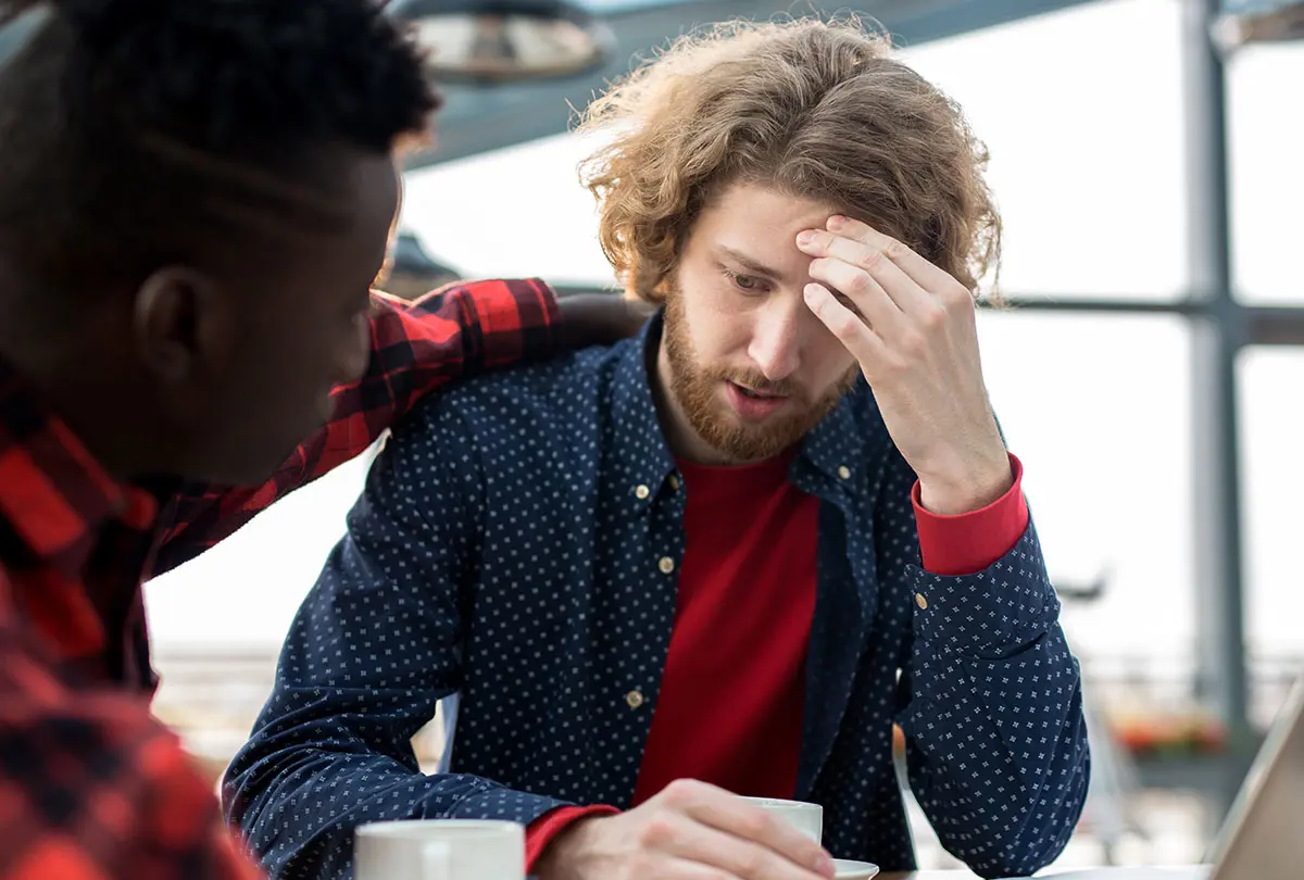 Two men sitting at a table, visibly distressed, with one resting his head in his hands.