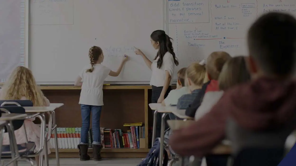 Teacher and student in a classroom standing at a whiteboard solving math problems.