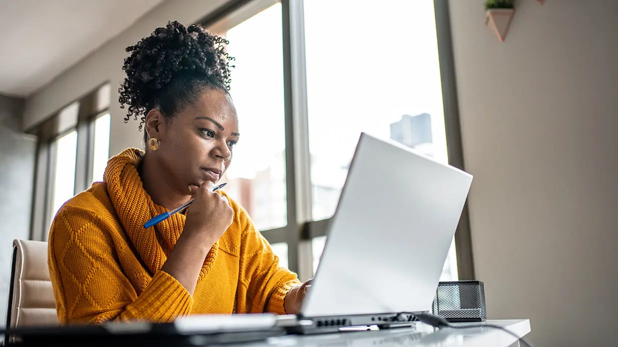 Woman in orange sweater holding a pen and looking at a laptop.